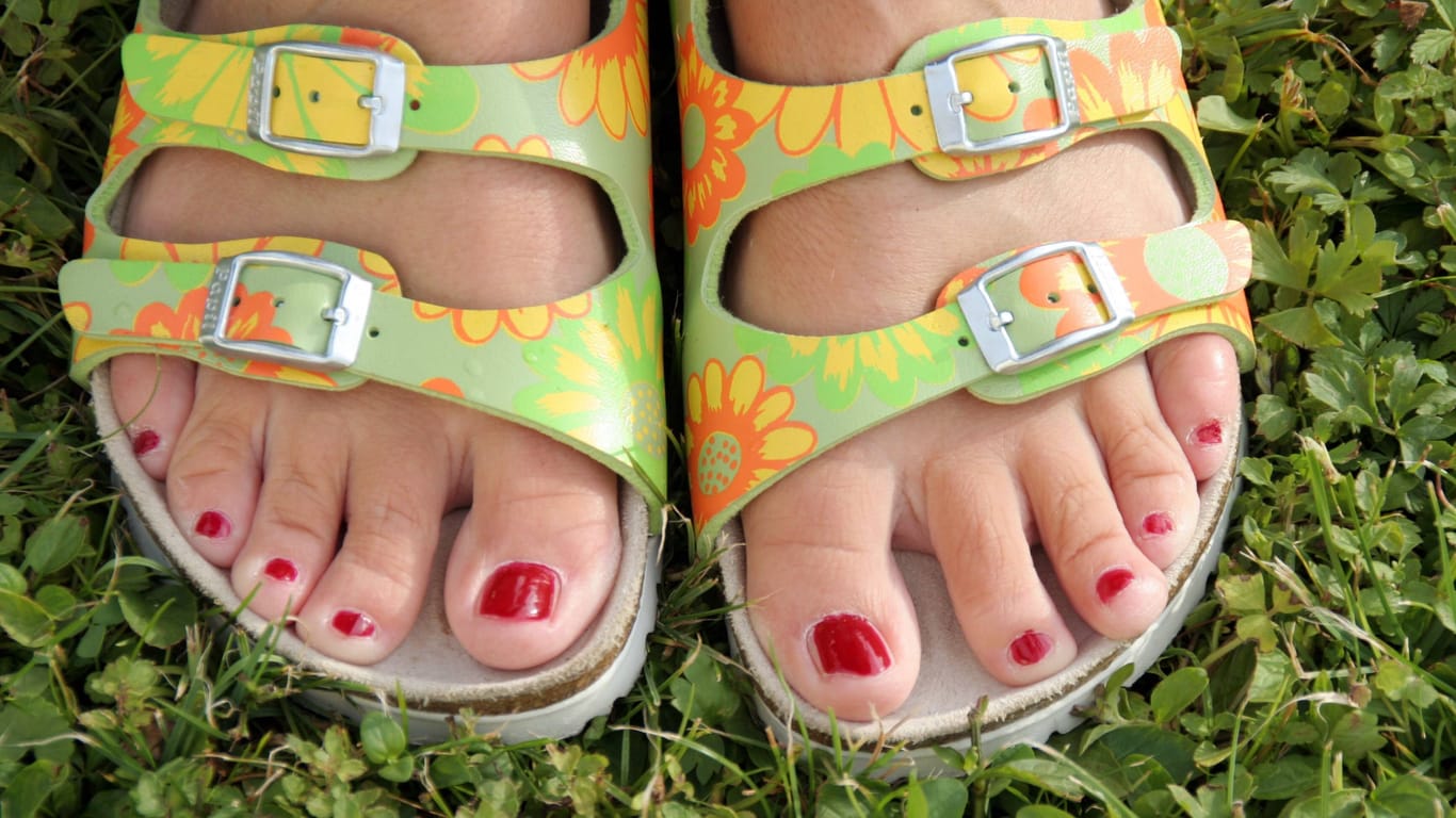 Frauenfüße in bunten Sandalen auf einer Wiese: Mit der richtigen Pflege halten Sommersandalen länger.