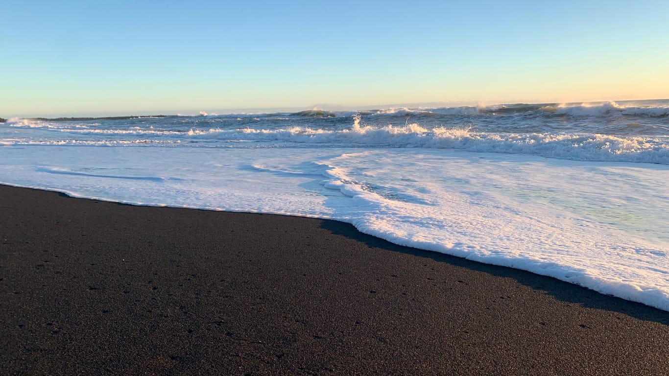 Die Sonne am Reynisfjara-Strand geht unter, schäumendes Atlantikwasser rauscht auf den schwarzen Sand.