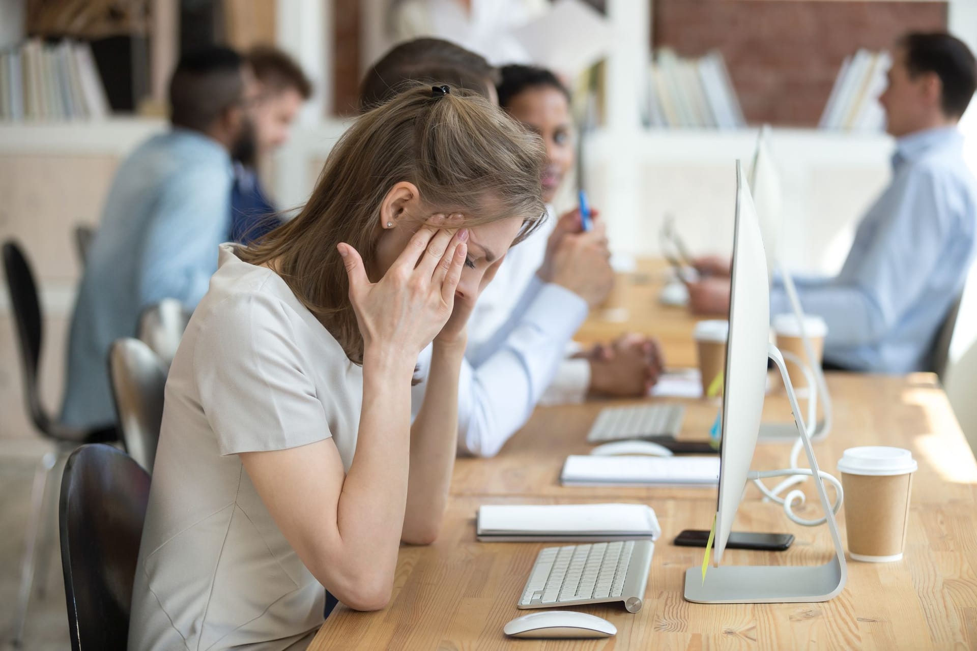 Eine Frau sitzt vor einem Computer im Büro. Sie hält sich die Schläfen und sieht müde aus.