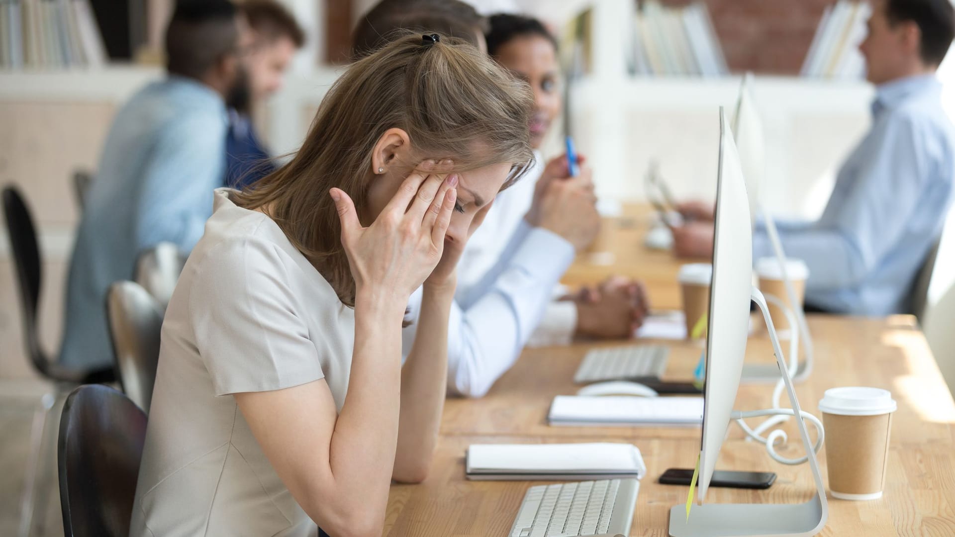 Eine Frau sitzt vor einem Computer im Büro. Sie hält sich die Schläfen und sieht müde aus.