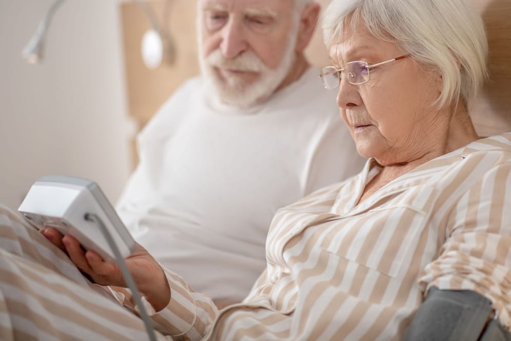 Ältere Frau misst neben einem Mann im Bett liegend ihren Blutdruck