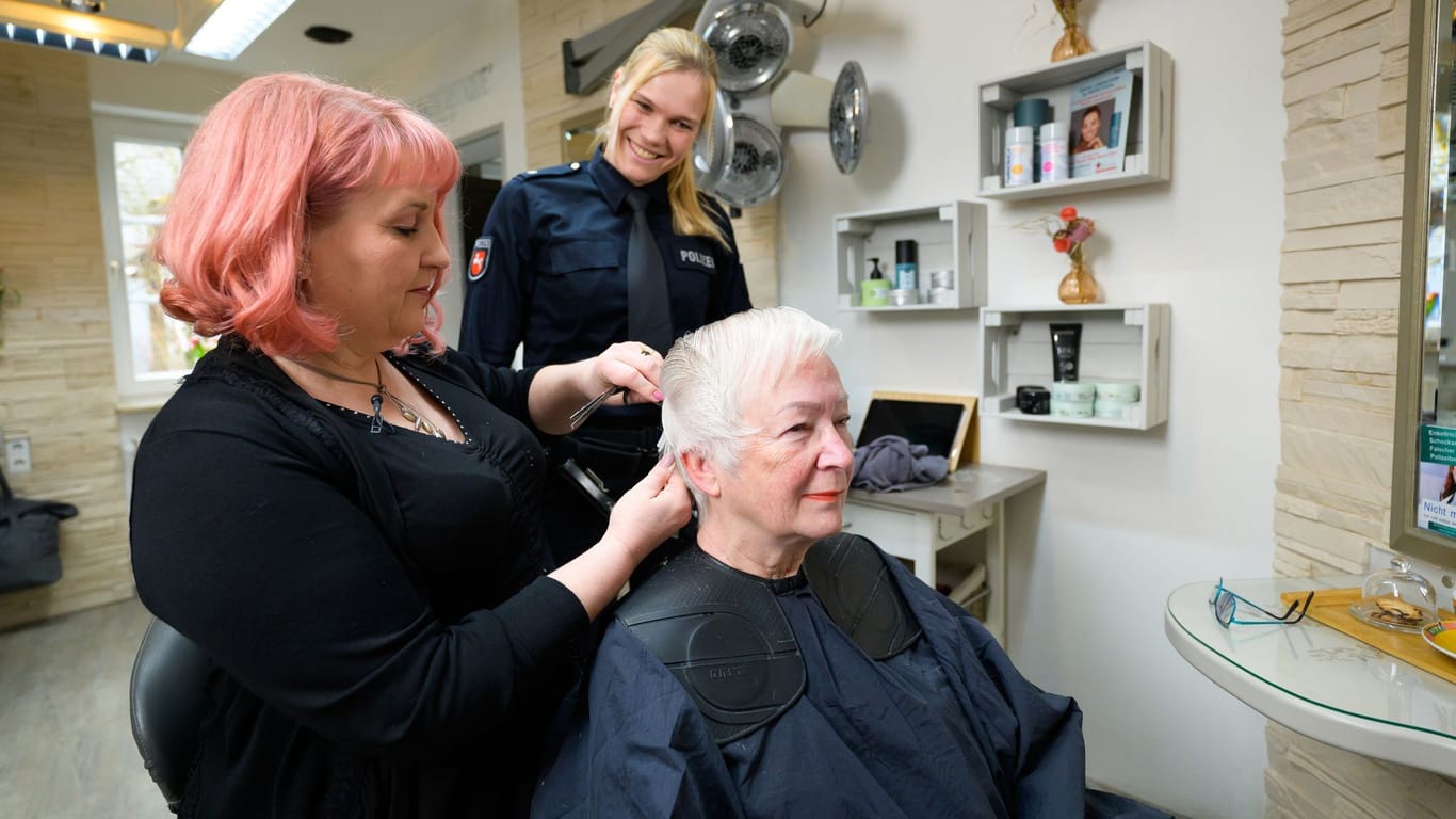 Claudia Schmidt (l), Friseurmeisterin, schneidet Doris Deuhs-Wagener (r) die Haare. Stella Giese von der Polizei schaut ihr dabei über die Schulter.
