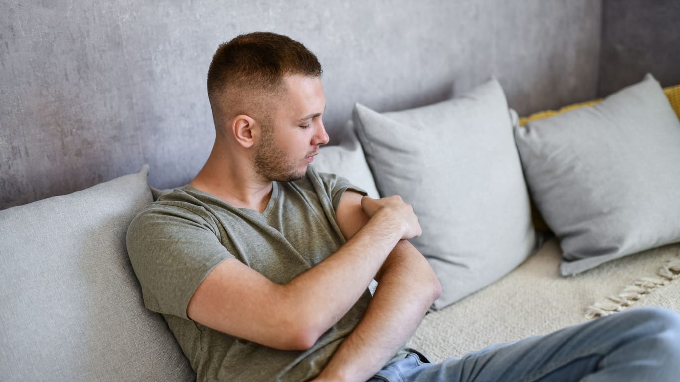 Ein Mann sitzt auf dem Sofa und betrachtet seinen linken Oberarm.