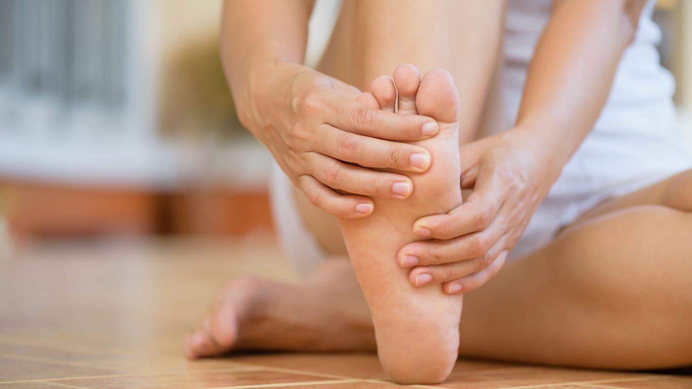 Auf dem Boden sitzende Frau umschließt ihren rechten Fuß mit den Händen: Eine Plantarfasziitis lässt sich mit verschiedenen Maßnahmen behandeln.
