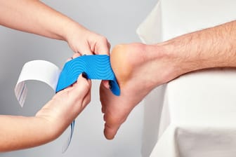Eine Therapeutin befestigt Kinesiotape an der Ferse eines Patienten: Bei einer Plantarfasziitis den Fuß zu tapen, soll die Behandlung unterstützen.