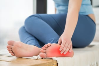 Eine Frau berührt ihren linken Fuß: Hält die Überlastung an, können sich die Schmerzen bei einer Plantarfasziitis verschlimmern.