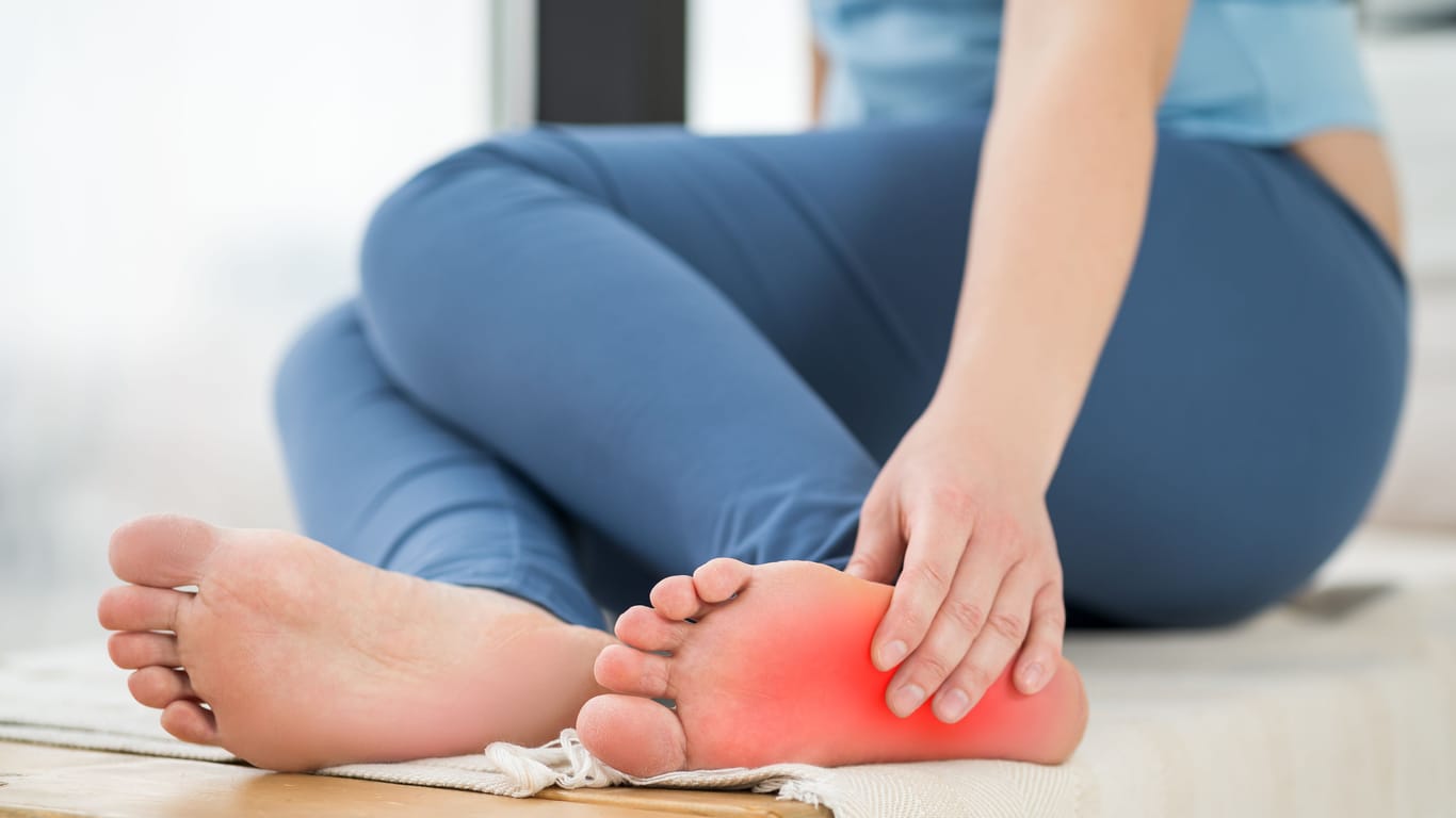 Eine Frau berührt ihren linken Fuß: Hält die Überlastung an, können sich die Schmerzen bei einer Plantarfasziitis verschlimmern.