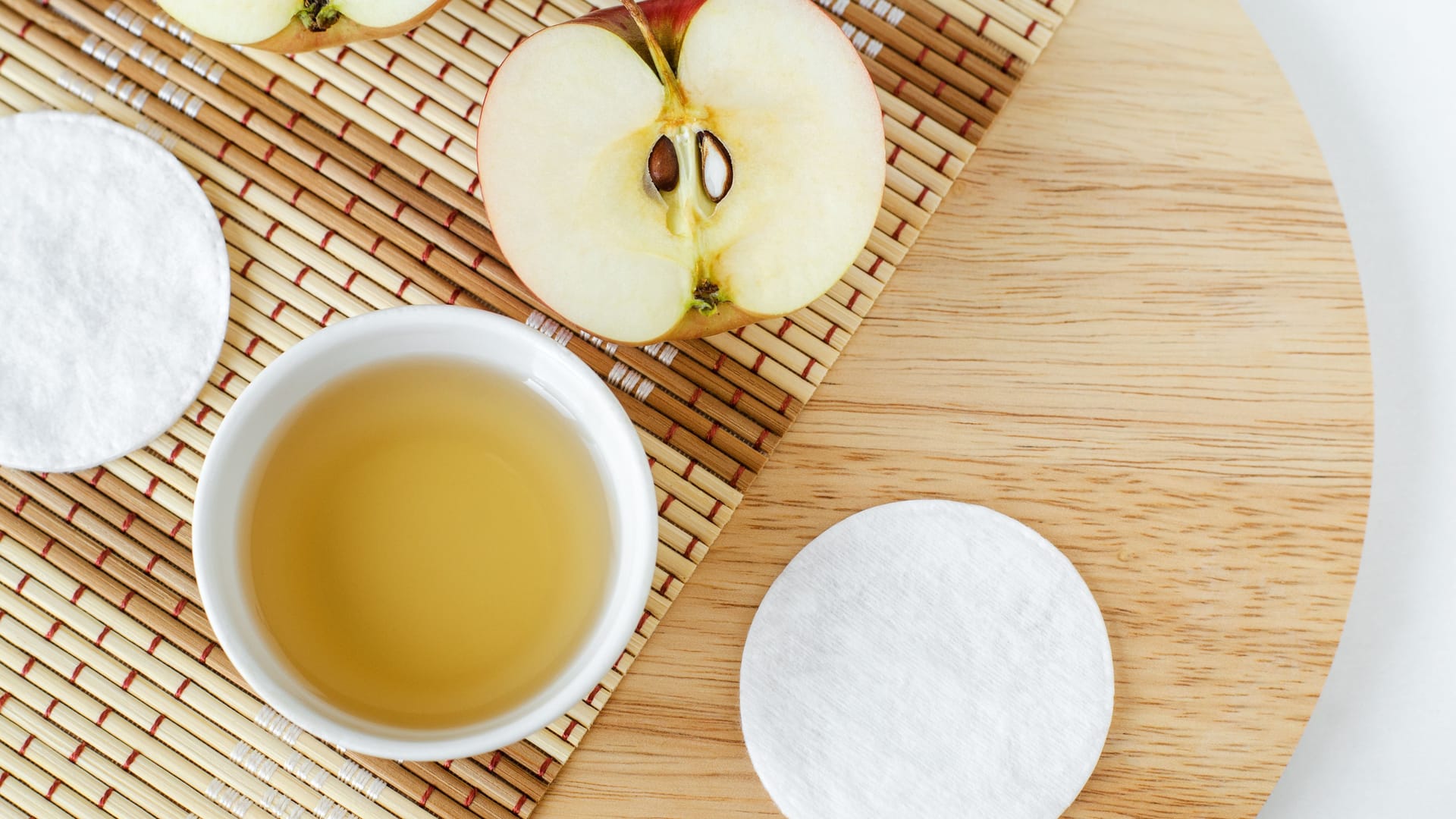 Ein Gefäß mit Apfelessig, ein Wattepad und ein Apfel liegen auf einem Holzbrett.