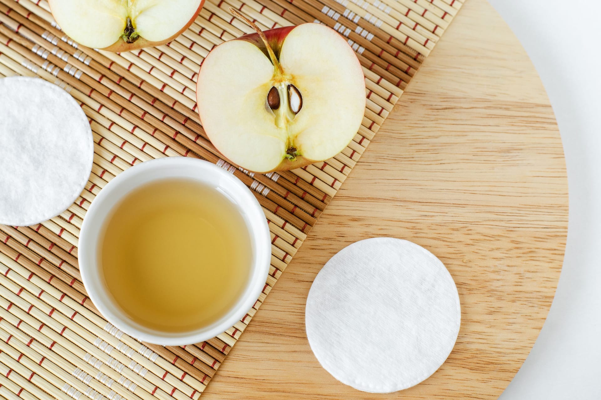 Ein Gefäß mit Apfelessig, ein Wattepad und ein Apfel liegen auf einem Holzbrett.