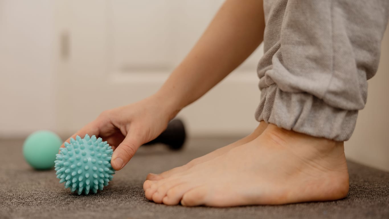 Eine Frau greift nach einem Igelball vor ihren Füßen: Den Fuß über einen Igelball, eine kleine Faszienrolle oder eine gekühlte Wasserflasche zu rollen, kann sich bei einer Plantarfasziitis günstig auswirken.
