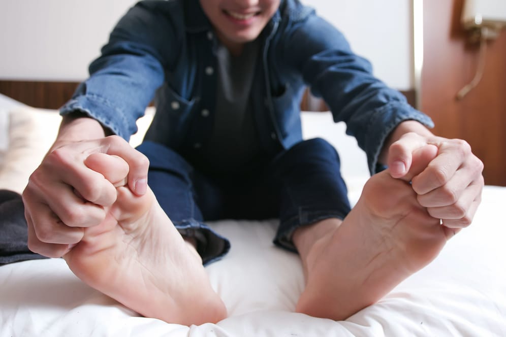 Ein Mann sitzt auf dem Bett und umklammert seine Zehen.