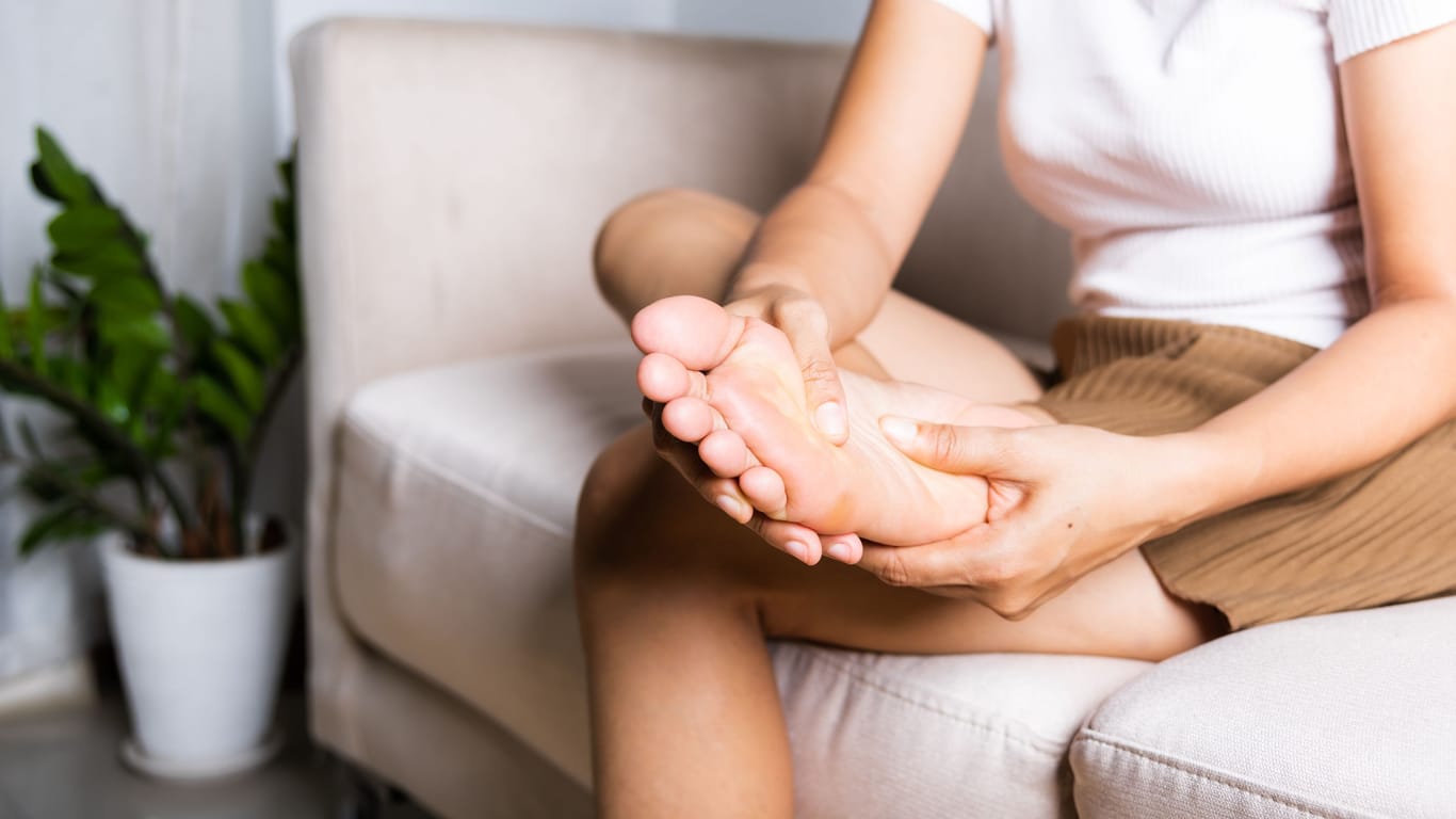 Eine Frau auf einem Sofa hält ihren rechten Fuß in den Händen: Gegen die Fußschmerzen bei einer Plantarfasziitis kann eine Massage infrage kommen.