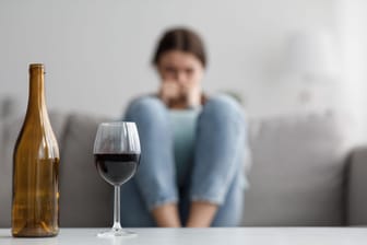 Eine Person sitzt auf einem Sofa, vor ihr ein Glas Rotwein und eine leere Flasche: Alkoholkonsum birgt viele Risiken.