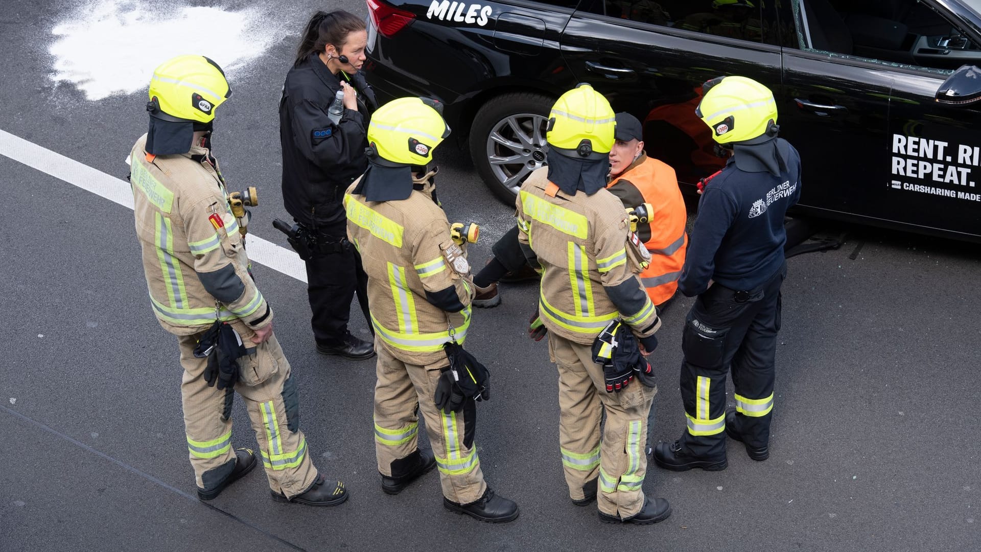 Feuerwehrleute und eine Polizistin stehen neben einem Aktivisten: Er hatte sich unter einem Auto auf die Fahrbahn geklebt.