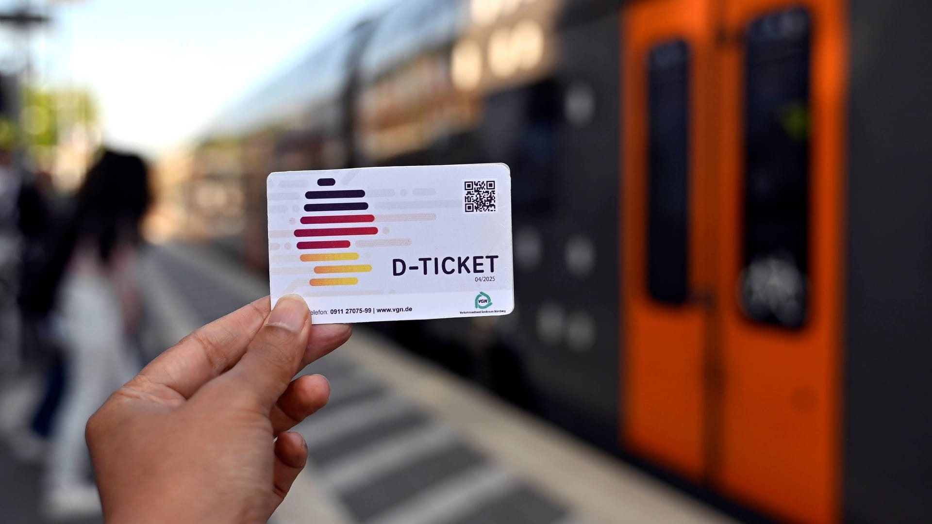 Jemand hält sein Deutschland-Ticket neben einer Bahn: Das Deutschland-Ticket gilt als der Nachfolger des 9-Euro-Tickets.