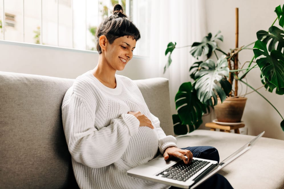 Schwangere sitzt mit Laptop auf dem Sofa