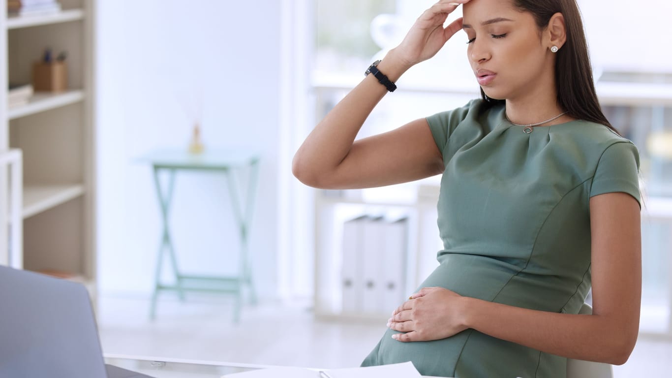 Schwangere Frau am Schreibtisch fasst sich wegen Unwohlseins mit einer Hand an die Stirn
