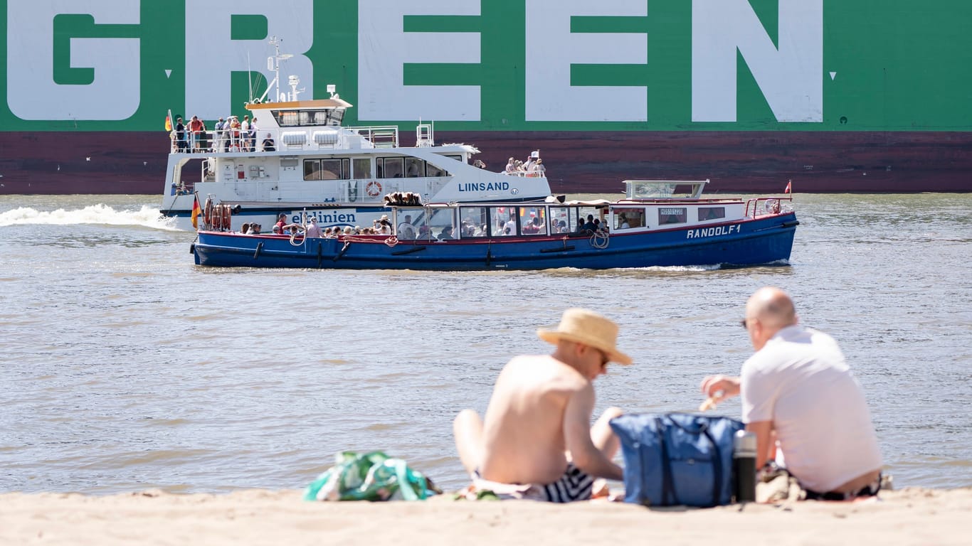 Zwei Menschen genießen die Sonne am Elbstrand in Hamburg (Symbolbild): Im Sand sitzend lassen sich von dort die großen Containerschiffe beobachten.