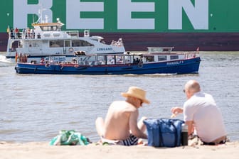Zwei Menschen genießen die Sonne am Elbstrand in Hamburg (Symbolbild): Im Sand sitzend lassen sich von dort die großen Containerschiffe beobachten.