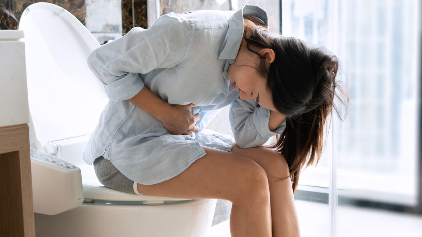 Frau mit Bauchschmerzen sitzt auf der Toilette: Eine EHEC-Infektion geht oft mit wässrigem Durchfall und weiteren Beschwerden einher.