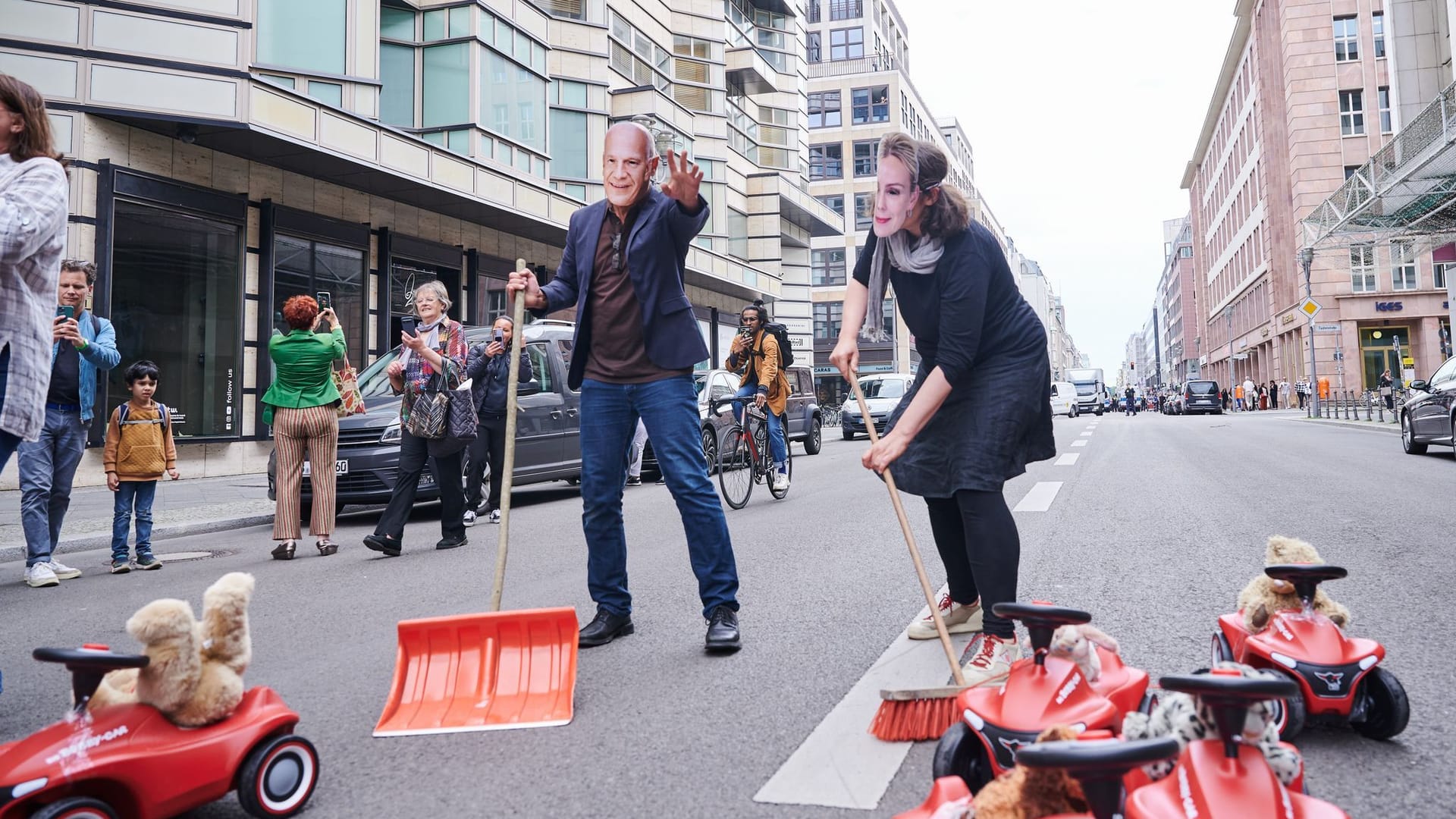 Ein Demonstrant mit einer "Kai-Wegner-Maske" und eine Demonstrantin mit "Manja-Schreiner-Maske": Sie kehren symbolisch auf der Friedrichstraße die Bobby-Cars aus dem Weg.