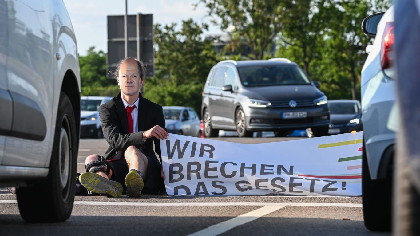 Ein Mitglied der "Letzte Generation" nimmt an einer Sitzblockade an der Autobahnabfahrt am Elbepark in Dredsen teil: Sie tragen dabei eine Maske mit dem Porträt von Bundeskanzler Scholz.
