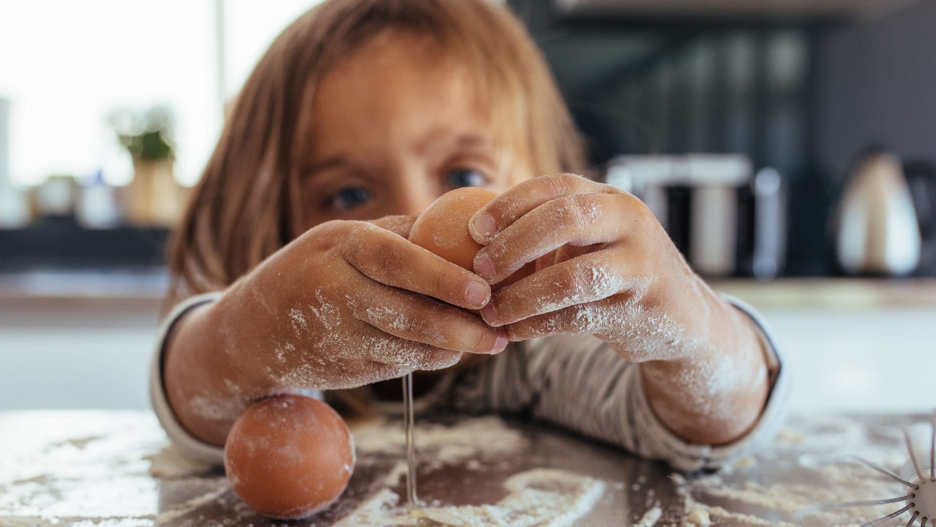 Kind schlägt ein Ei über bemehlter Küchenarbeitsfläche auf Kind schlägt ein Ei über bemehlter Küchenarbeitsfläche auf