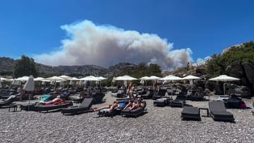 Paradoxes Foto von der Insel Lindos: Menschen sonnen sich am Strand, während am Himmel Rauchwolken von den Bränden auf Rhodos aufsteigen.