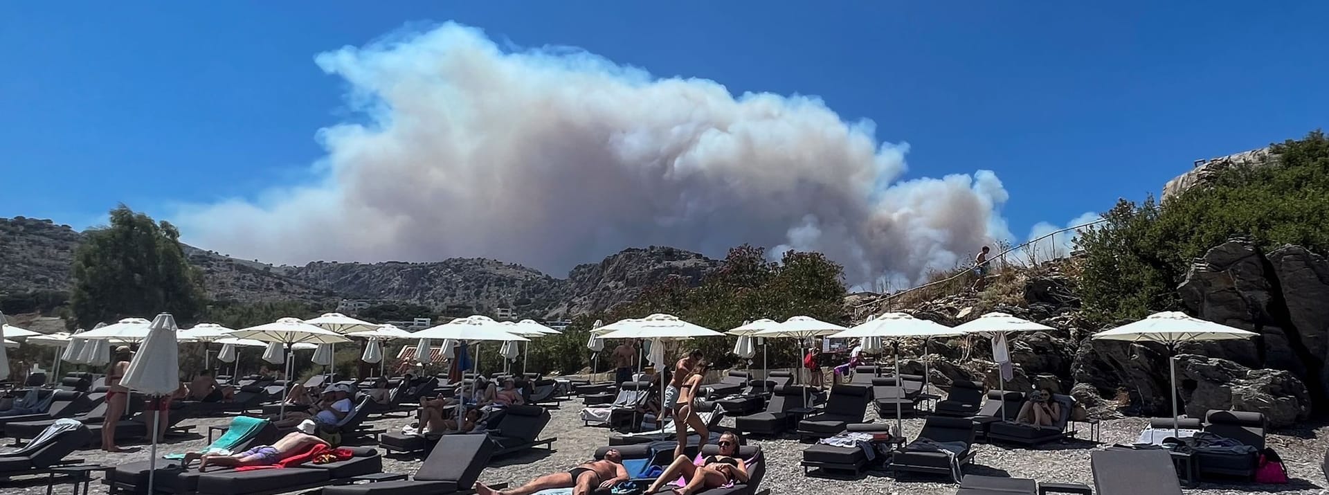 Paradoxes Foto von der Insel Lindos: Menschen sonnen sich am Strand, während am Himmel Rauchwolken von den Bränden auf Rhodos aufsteigen.