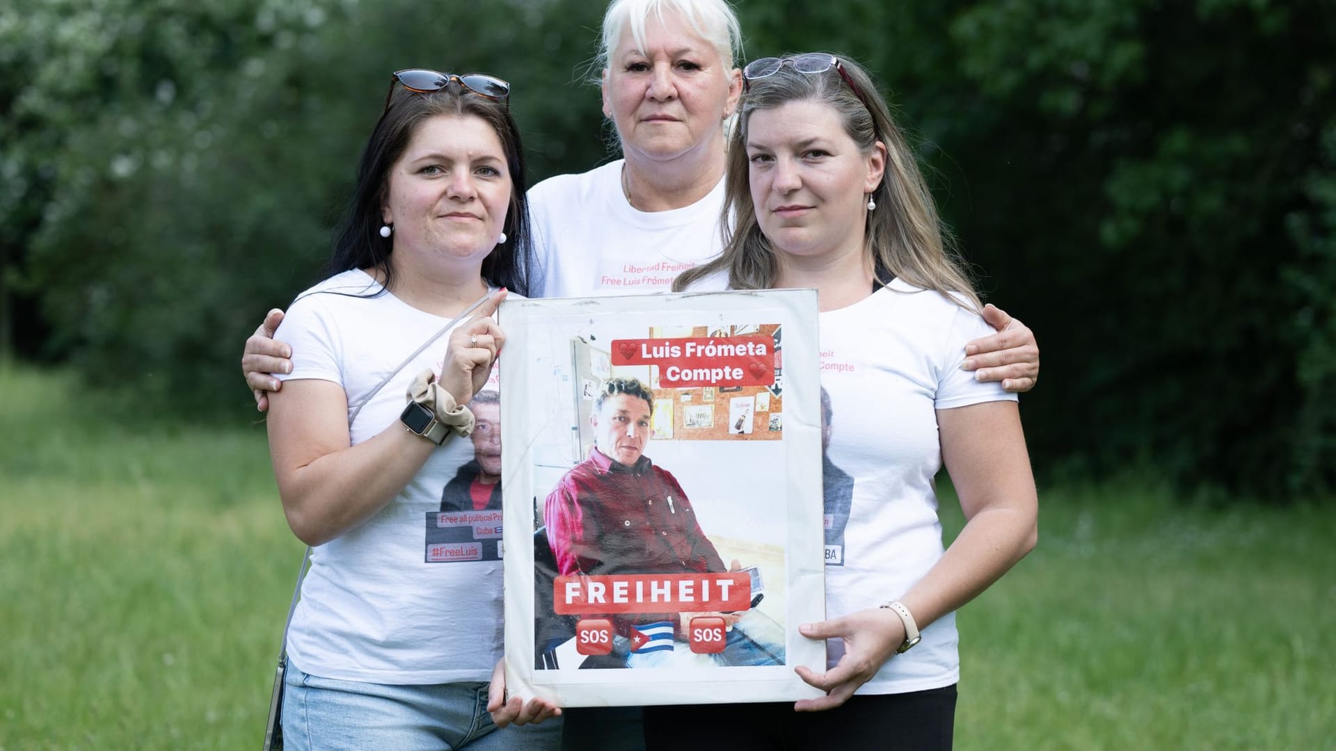 Maria (l) und Janie (r) Frometa Compte, Töchter des inhaftierten Deutsch-Kubaners Luis Frometa Compte, und Silke Frometa Compte, Ehefrau, stehen am Rande einer Kundgebung im Alaunpark und halten ein Foto von Luis Frometa Compte. Der Dresdner Frómeta Compte war im Juli 2021 auf Familienbesuch in Havanna gewesen, als es in dem sozialistischen Karibikstaat in vielen Städten zu Demonstrationen für Freiheit und gegen Misswirtschaft kam. Frómeta Compte wurde nach Angaben seiner Familie festgenommen, weil er mit dem Handy einen der Proteste filmte. Er wurde wegen Aufruhr verurteilt, eine ursprüngliche 25-jährige Haftstrafe wurde auf 15 Jahre reduziert.