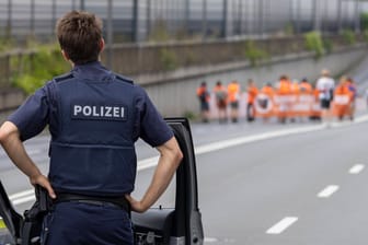 Ein Polizist blickt Mitte August auf Mitglieder der "Letzten Generation", die die B19 in Würzburg blockieren Auf ihrem Transparent steht: "Schöne lange Welt, wie lange noch?"