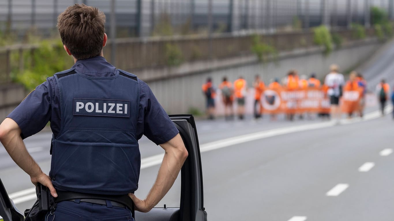 Ein Polizist blickt Mitte August auf Mitglieder der "Letzten Generation", die die B19 in Würzburg blockieren Auf ihrem Transparent steht: "Schöne lange Welt, wie lange noch?"