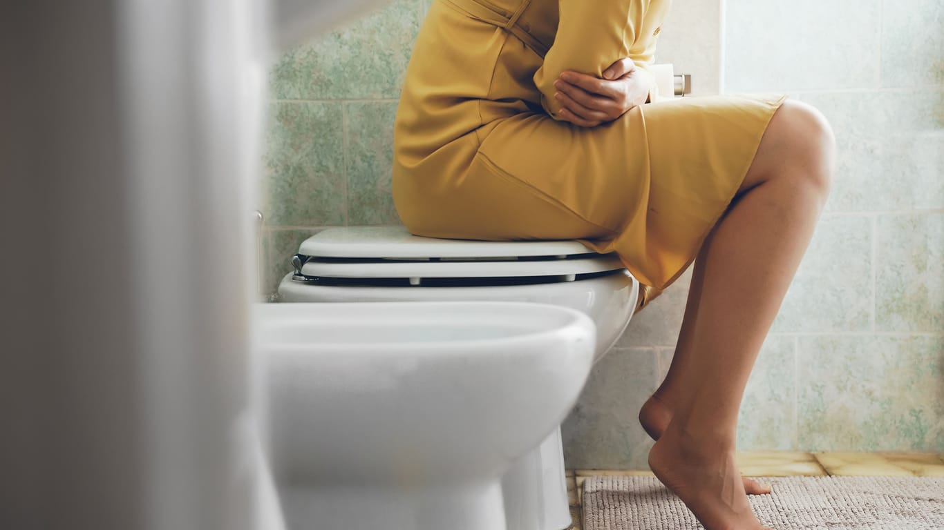 Frau mit Bauchschmerzen auf Toilette: Nach einer Fernreise kann es öfters einmal zu Durchfall kommen, meist nur für wenige Tage. In manchen Fällen halten die Beschwerden nach dem Urlaub aber länger an.