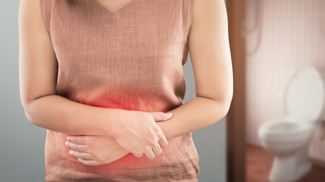 Eine Frau verschränkt die Arme vor dem Bauch, im Hintergrund sieht man eine Toilette: Mit Campylobacter verkeimte Lebensmittel sind häufig Ursache von Durchfall.