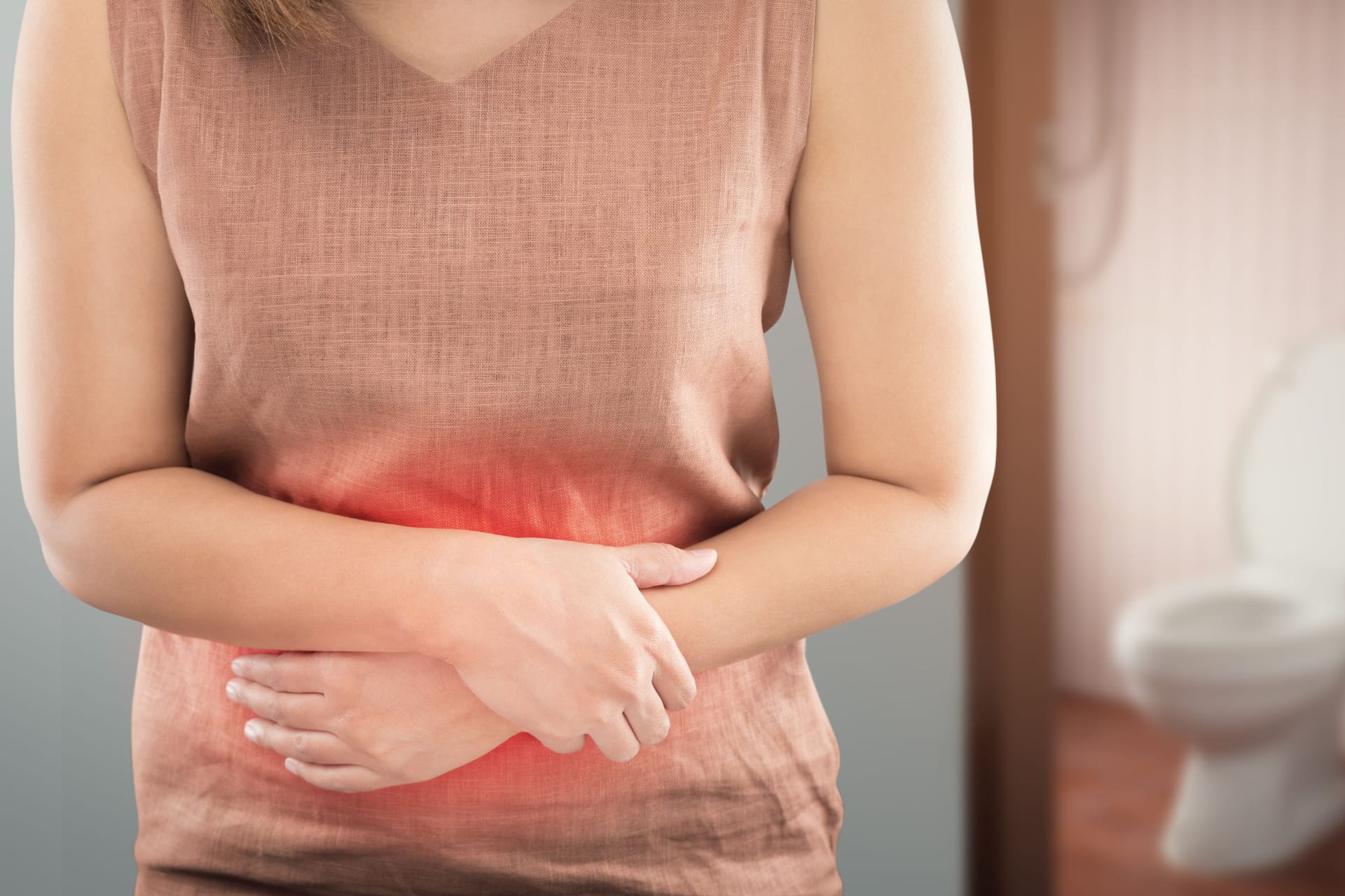 Eine Frau verschränkt die Arme vor dem Bauch, im Hintergrund sieht man eine Toilette: Mit Campylobacter verkeimte Lebensmittel sind häufig Ursache von Durchfall.