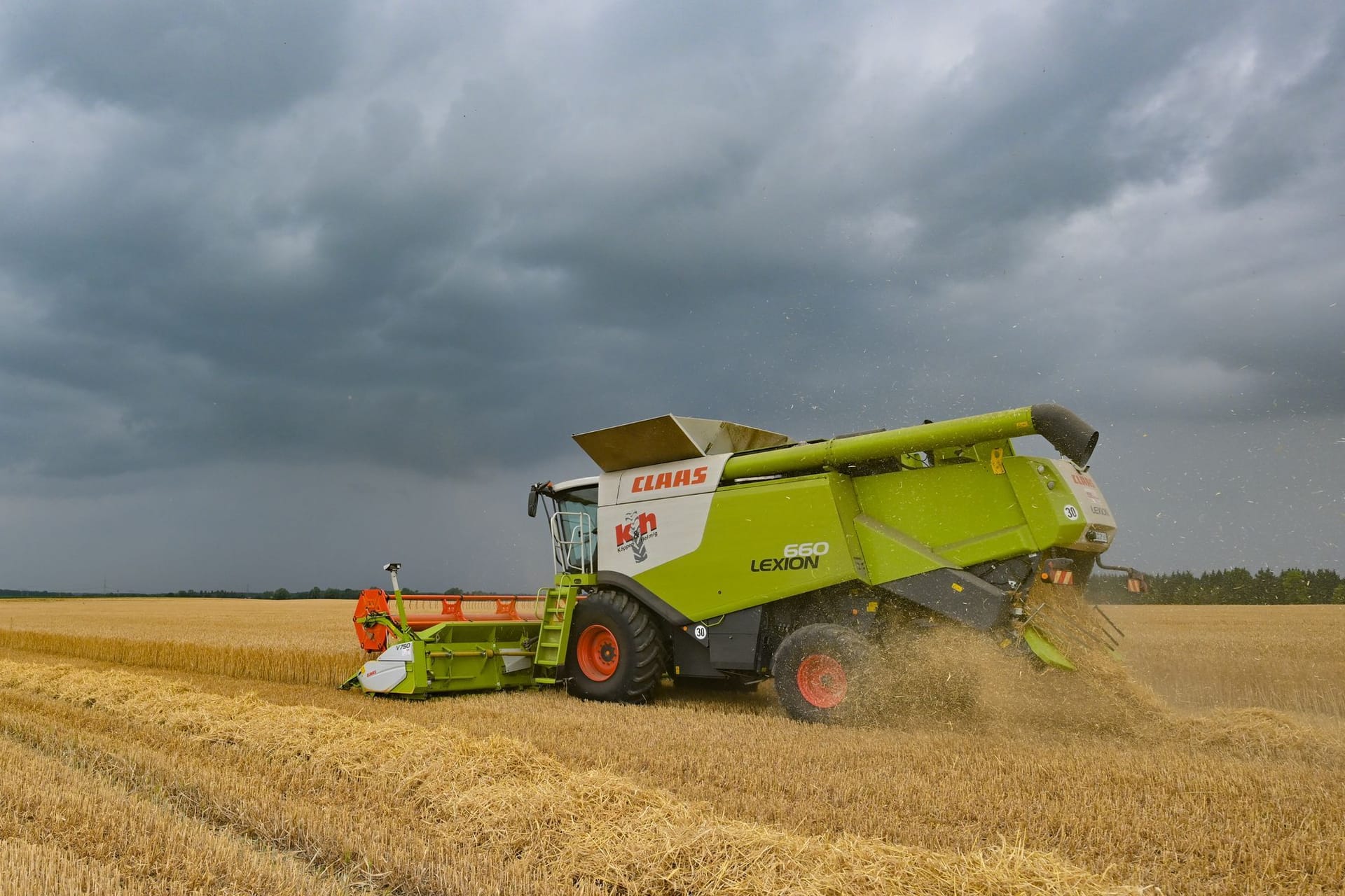 Dunkle Gewitterwolken ziehen über ein Feld, wo gerade ein Landwirt mit seinem Mähdrescher Wintergerste erntet.