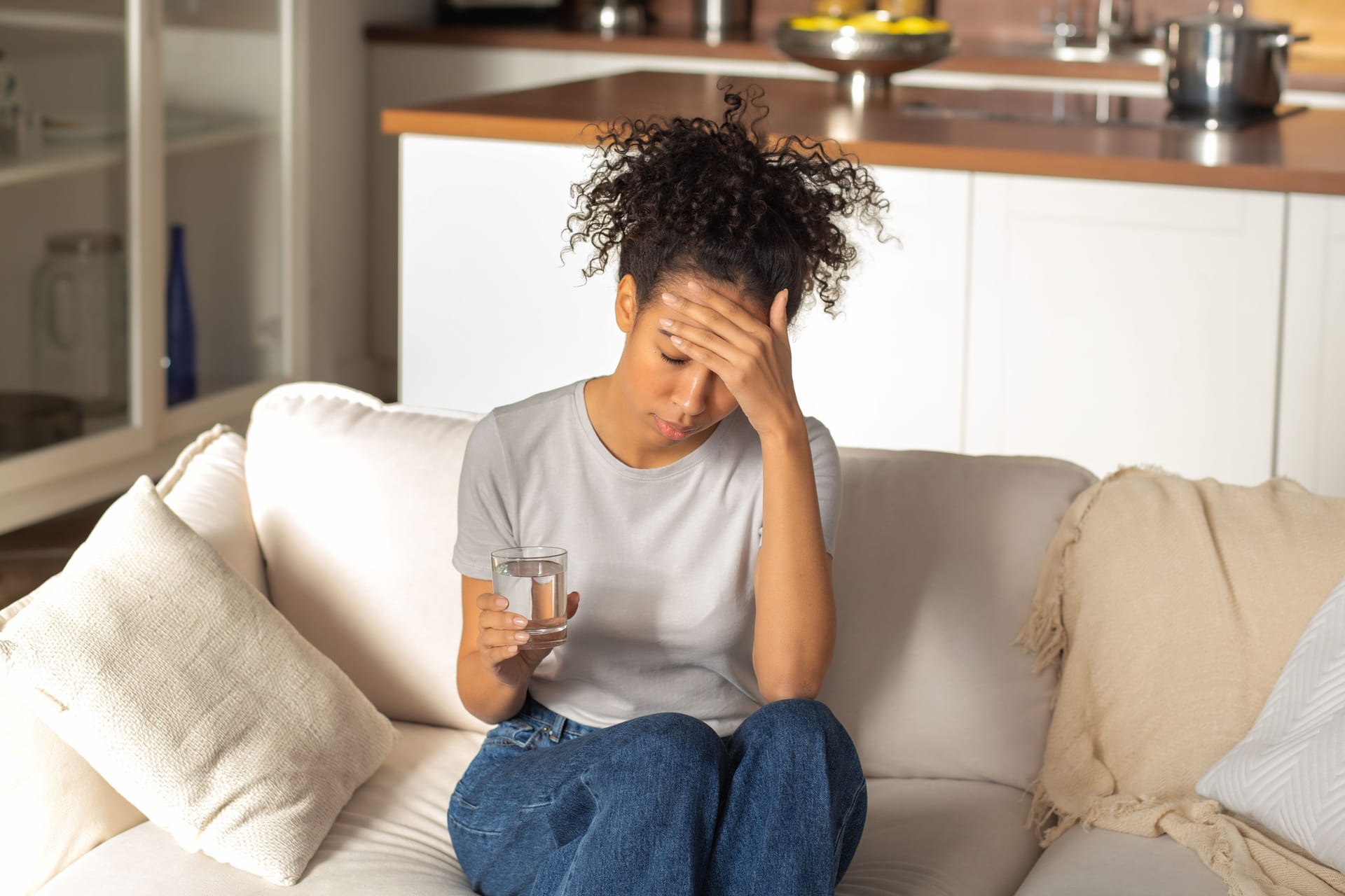 Eine Frau sitzt auf dem Sofa, hält sich den Kopf und trinkt ein Glas Wasser.