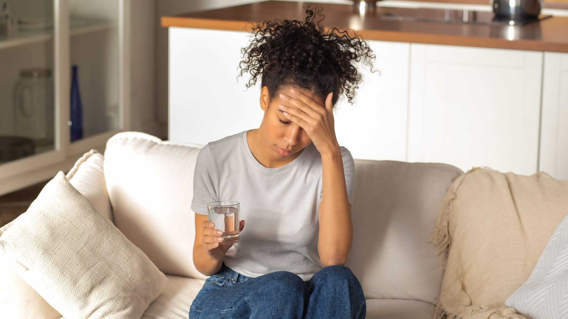 Eine Frau sitzt auf dem Sofa, hält sich den Kopf und trinkt ein Glas Wasser.