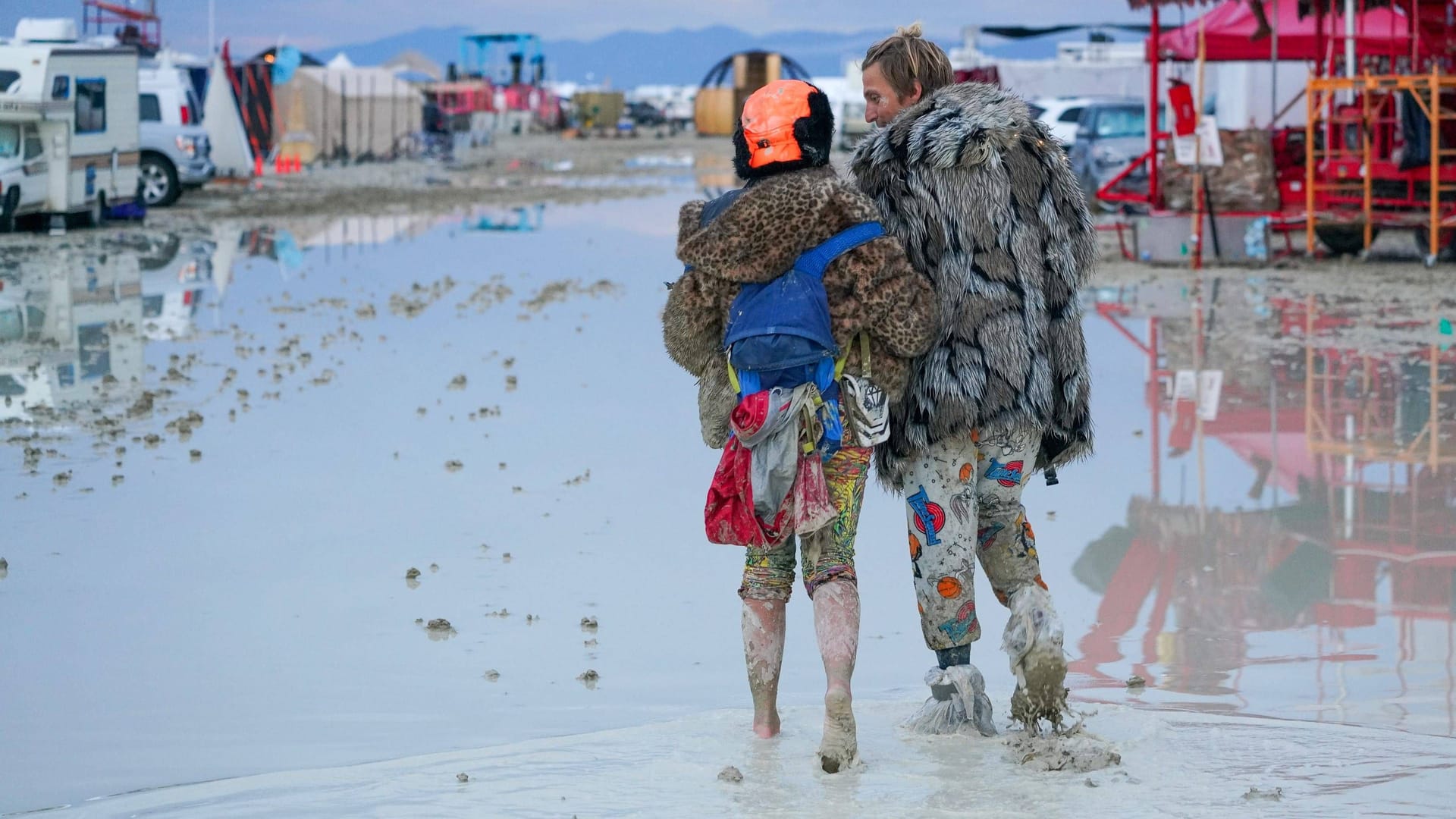 Zwei Festivalgänger streifen barfuß und mit Plastiktüten durch den Schlamm auf dem Gelände: Am Sonntagnachmittag Ortszeit soll es erneut heftig gewittern.