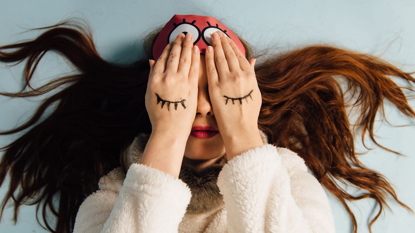 Frau mit Schlafmaske bedeckt die Augen mit den Händen: Wie viel wissen Sie über Schlaf? Testen Sie sich.