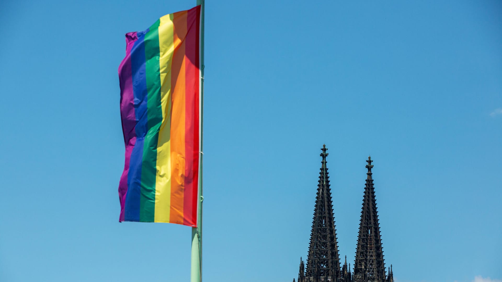 Regenbogenfahne mit Kölner Dom vor blauem Himmel: In Köln findet am Mittwoch ein Segnungsgottesdienst statt.