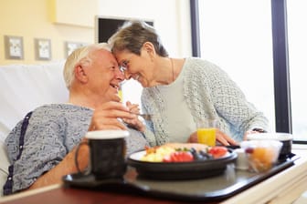 Ein Mann nimmt im Krankenhausbett eine Mahlzeit zu sich, während seine Frau zu Besuch ist.