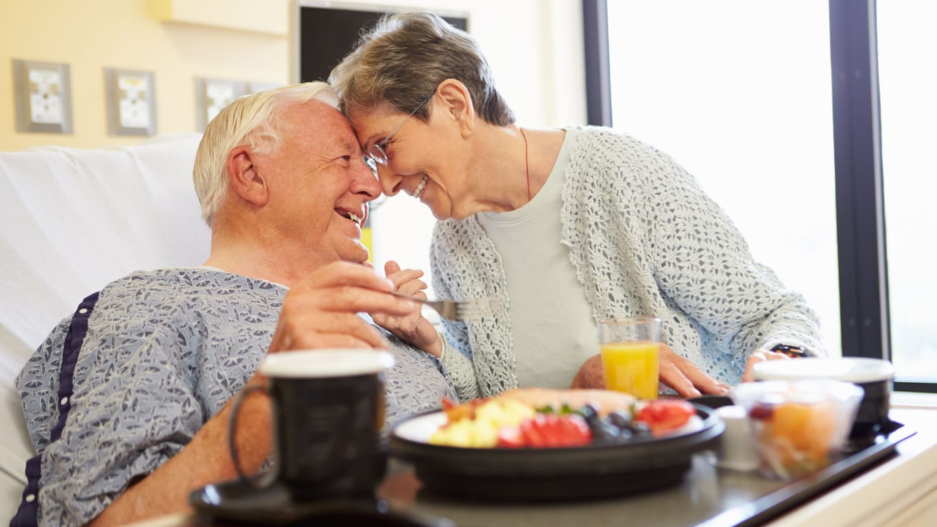 Ein Mann nimmt im Krankenhausbett eine Mahlzeit zu sich, während seine Frau zu Besuch ist.