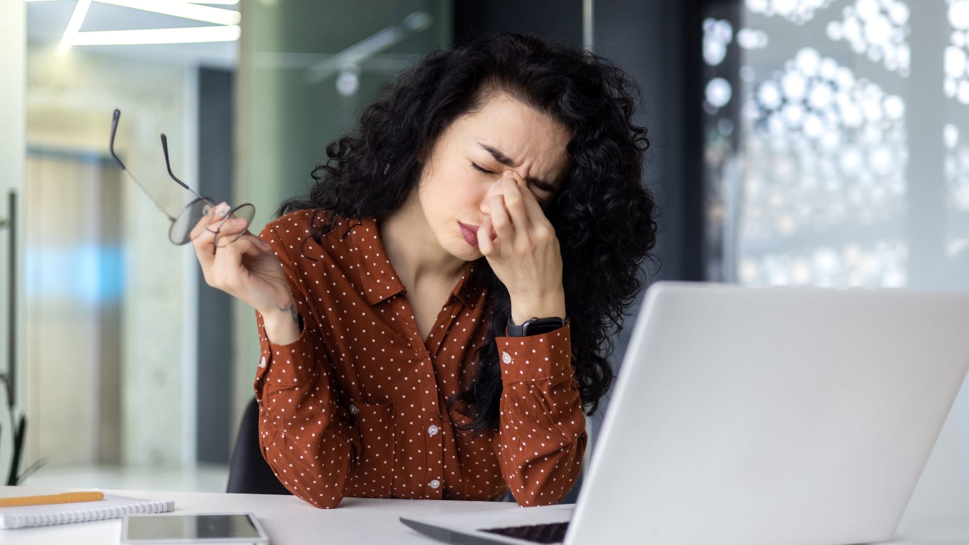 Eine Frau sitzt am Laptop und fasst sich mit der Hand an die Nasenwurzel.