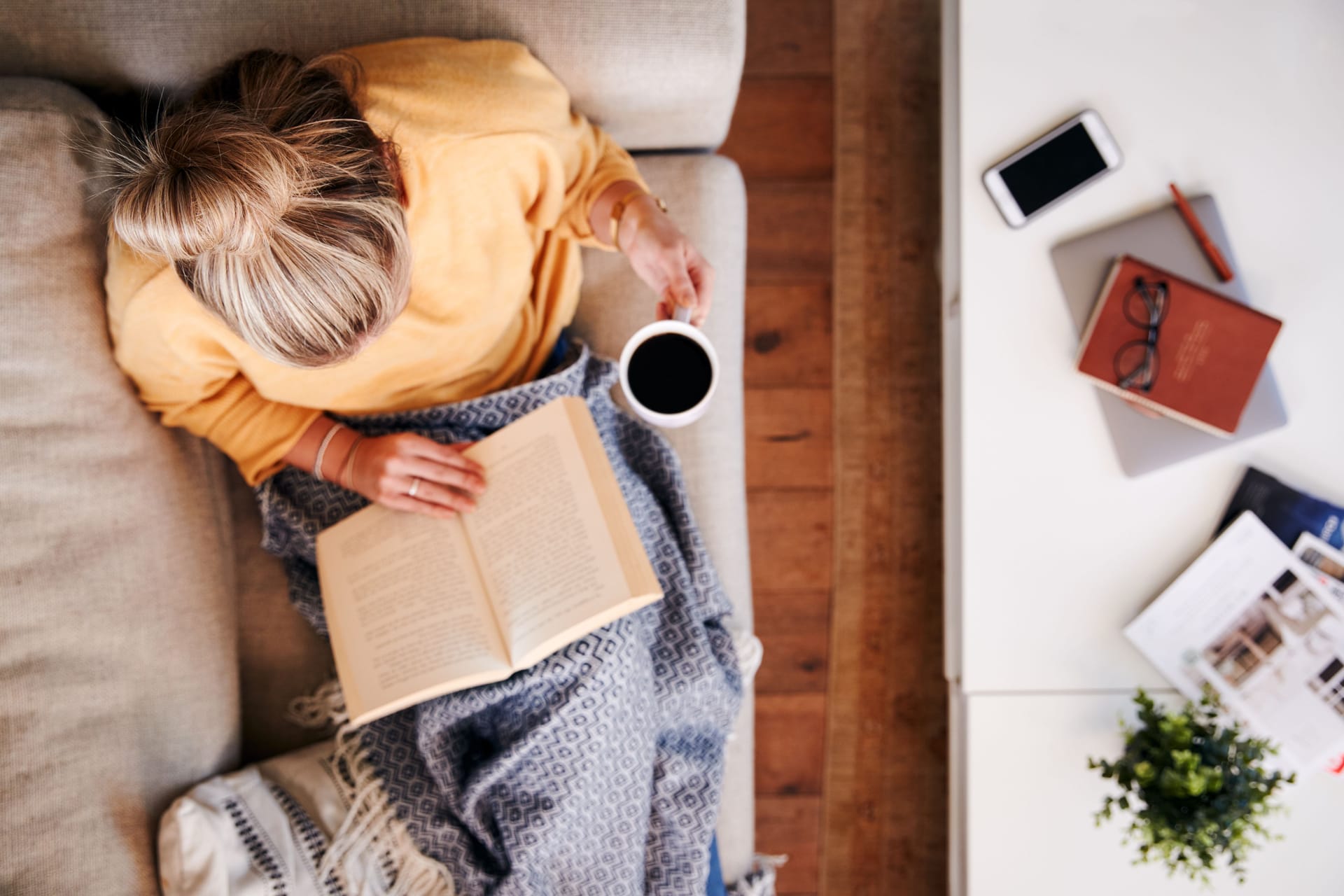 Eine Frau sitzt auf dem Sofa, liest und trinkt einen Kaffee.