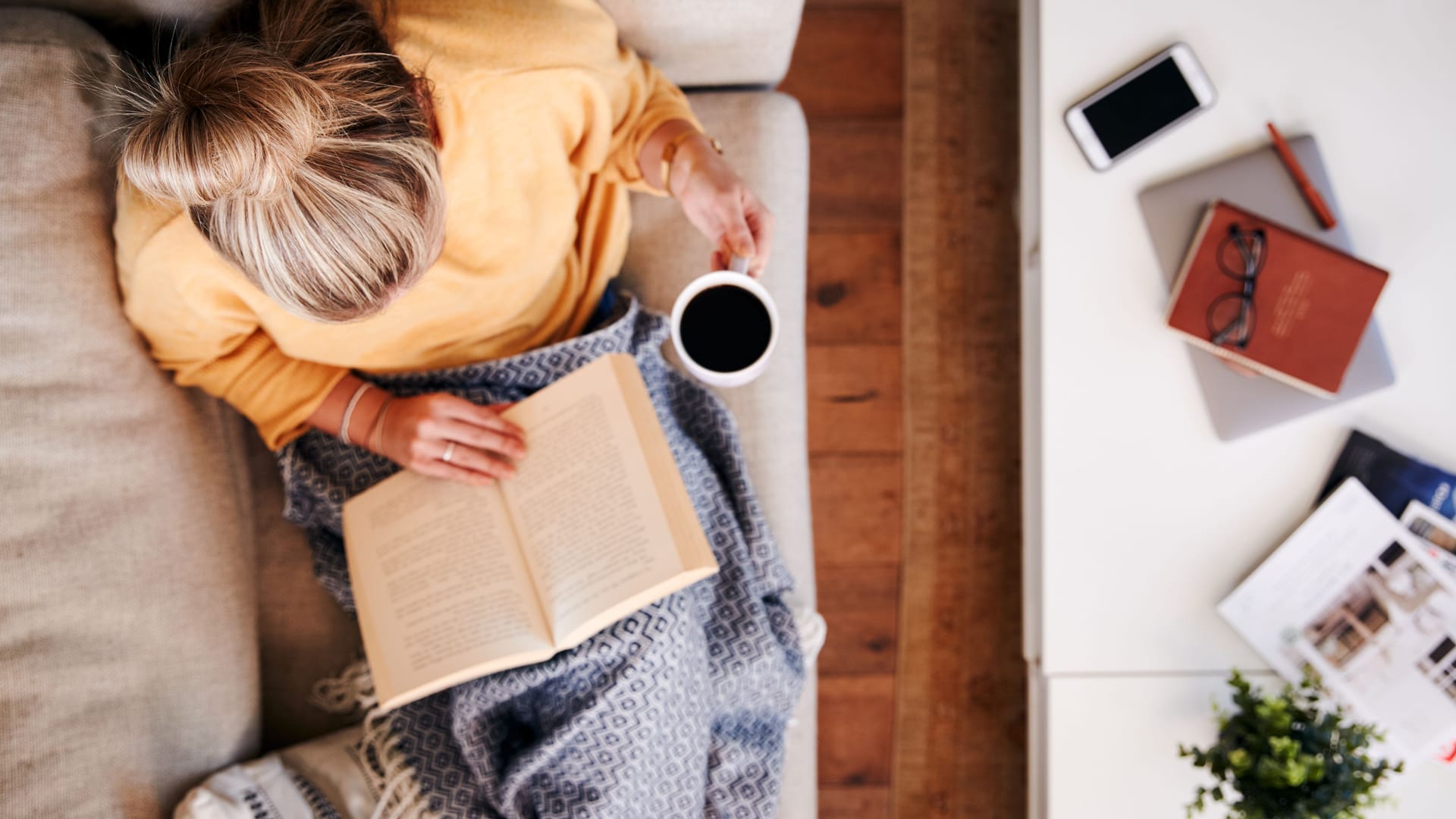 Eine Frau sitzt auf dem Sofa, liest und trinkt einen Kaffee.