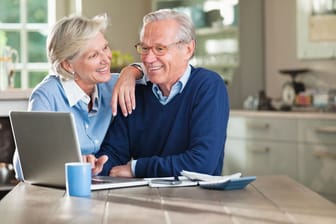 Eine Rentner-Ehepaar sitzt zu Hause am Tisch vor einem Laptop.