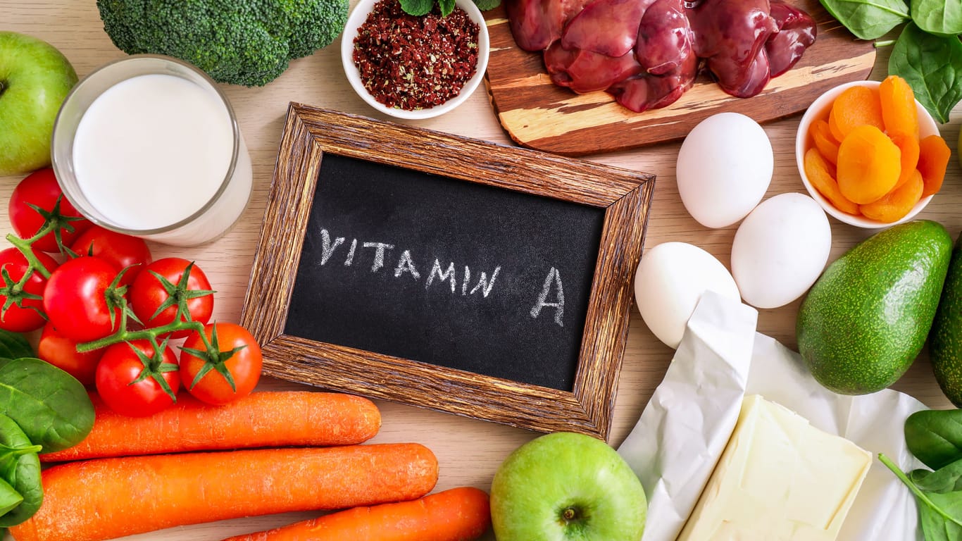 Man sieht Obst und Gemüse, Eier, Leber, Milch und Butter und eine Tafel mit der Aufschrift "Vitamin A": Um einem Vitamin-A-Mangel vorzubeugen und den Tagesbedarf zu decken, sollten täglich Lebensmittel mit Vitamin A oder Betacarotin verzehrt werden.