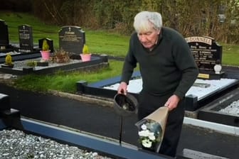 Alter Mann steht mit Blumen an einem Grab