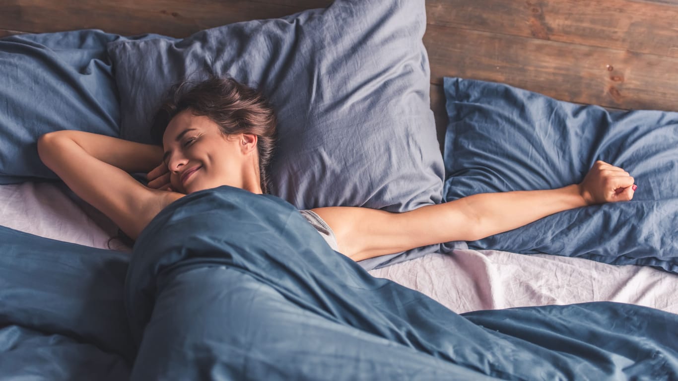 Eine Frau streckt sich entspannt im Bett: Wer leichte Einschlafprobleme hat, kann von der 4-7-8-Atmung wahrscheinlich profitieren.